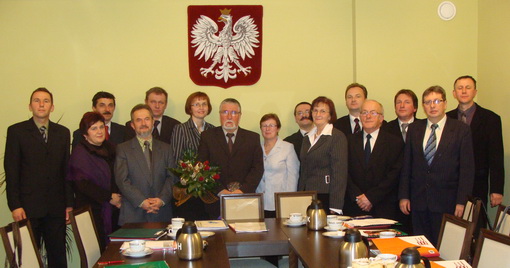 Radni Gminy Boron&oacute;w pozują do wsp&oacute;lnego zdjęcia w&nbsp;sali obrad. Stoją przy stole konferencyjnym, na&nbsp;kt&oacute;rym znajdują się dokumenty i&nbsp;naczynia. Na ścianie nad radnymi widoczne jest godło Polski.